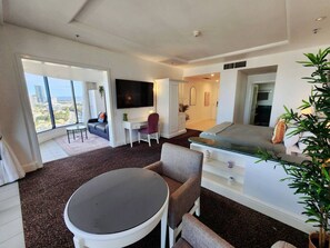 A modern hotel room with a large window, a flat-screen TV, a sofa, a dining table, and a small balcony.