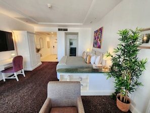 A hotel room with a bed, a desk, a chair, a potted plant, and a painting on the wall.