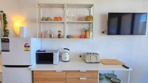 A compact kitchenette with a microwave, toaster, and a small refrigerator. There is a shelf above the counter with various items and a TV mounted on the wall. - Oaks Hotel - Deluxe Studio Apartment - Ocean Views (Surfers Paradise)