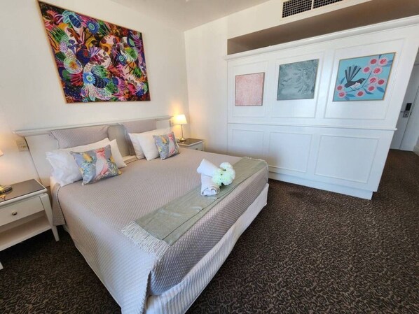 Una habitación de hotel con cama, mesitas de noche, un cuadro floral grande y un armario blanco decorado con obras de arte.