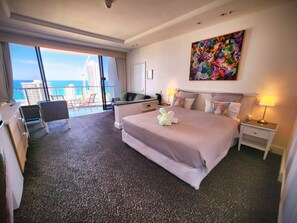 Habitación de hotel con una cama grande, un escritorio, una silla, un balcón con vista al mar y un cuadro colorido en la pared.