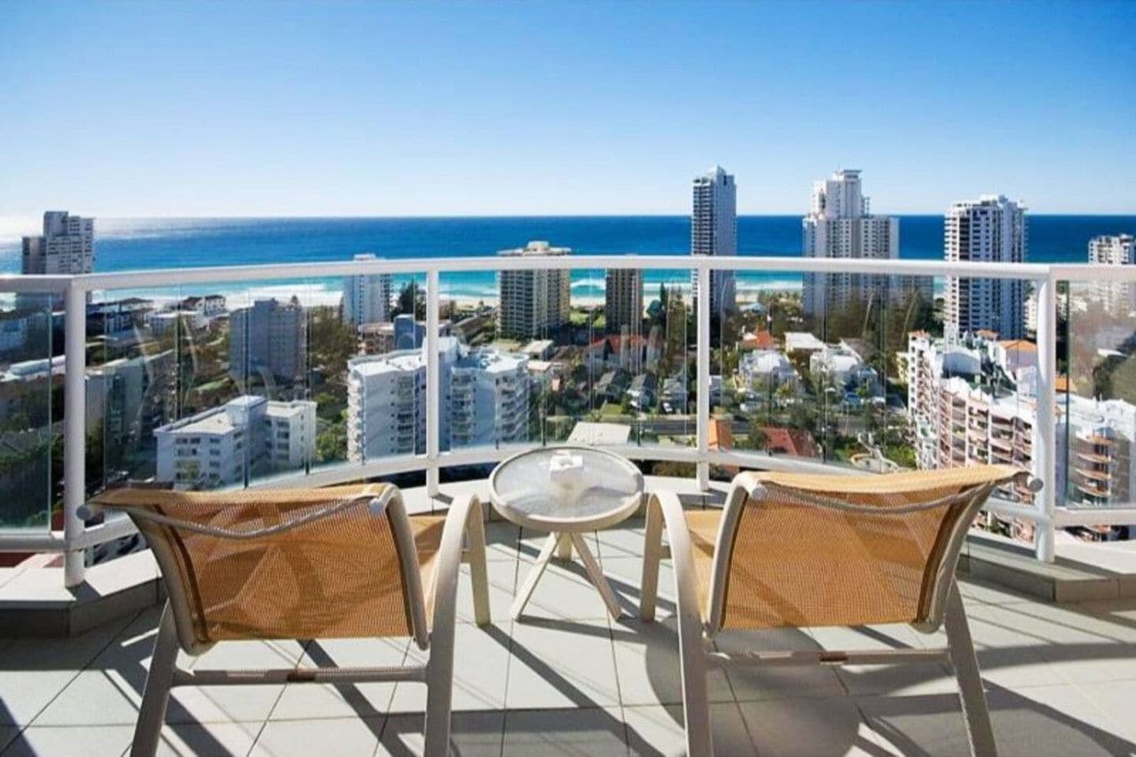 Een balkon met twee rieten stoelen en een kleine tafel, met uitzicht op een kustplaats en een helderblauwe lucht.