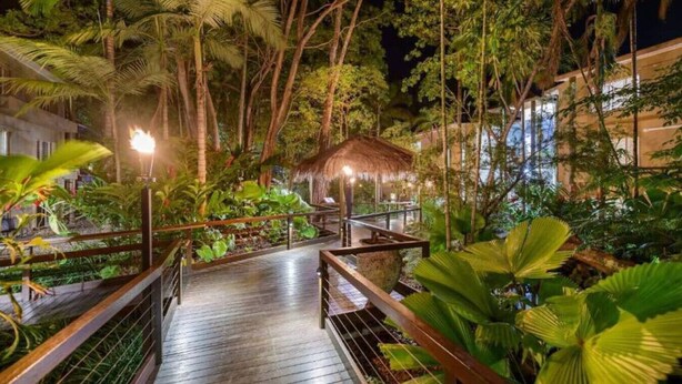 A wooden pathway leading through a lush garden with tropical plants and a thatched-roof structure.