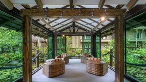 A covered outdoor seating area with wicker furniture and glass walls overlooking a lush garden.