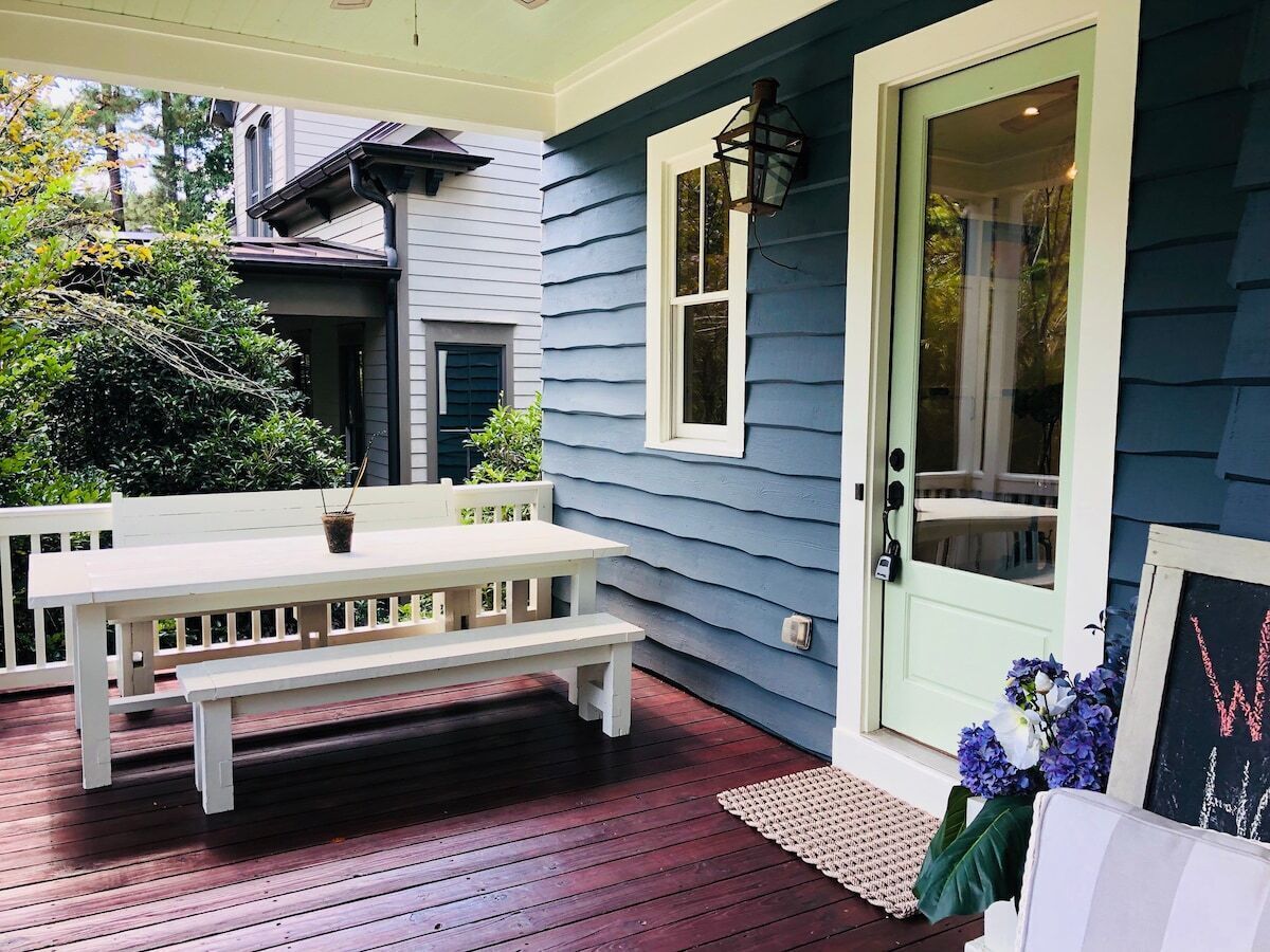 Une terrasse en bois avec un banc blanc et une table, une porte avec un cadre vert et un pot de fleurs.