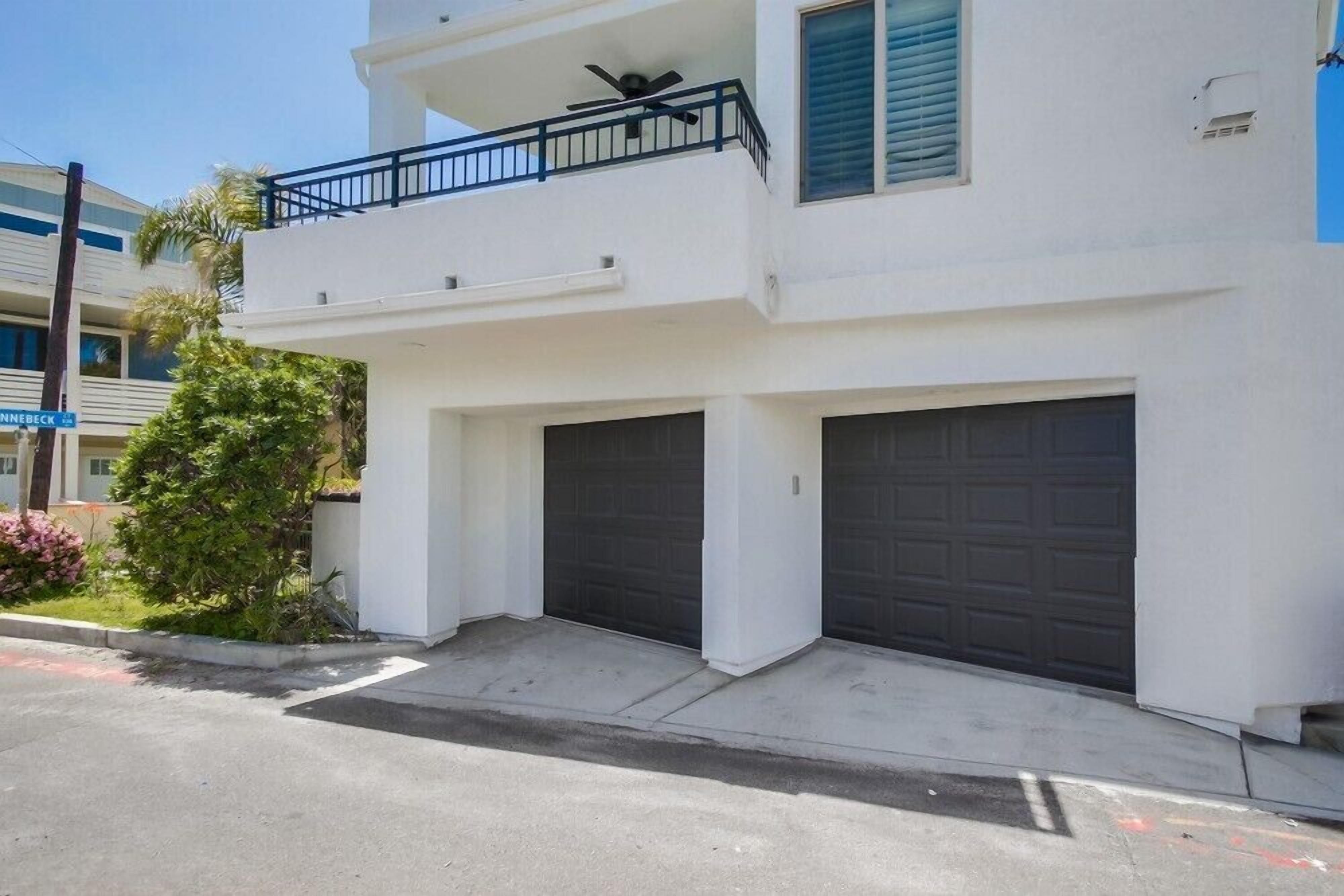A two-story residential building with a balcony, two garage doors, and a small front yard with a tree and flowers.