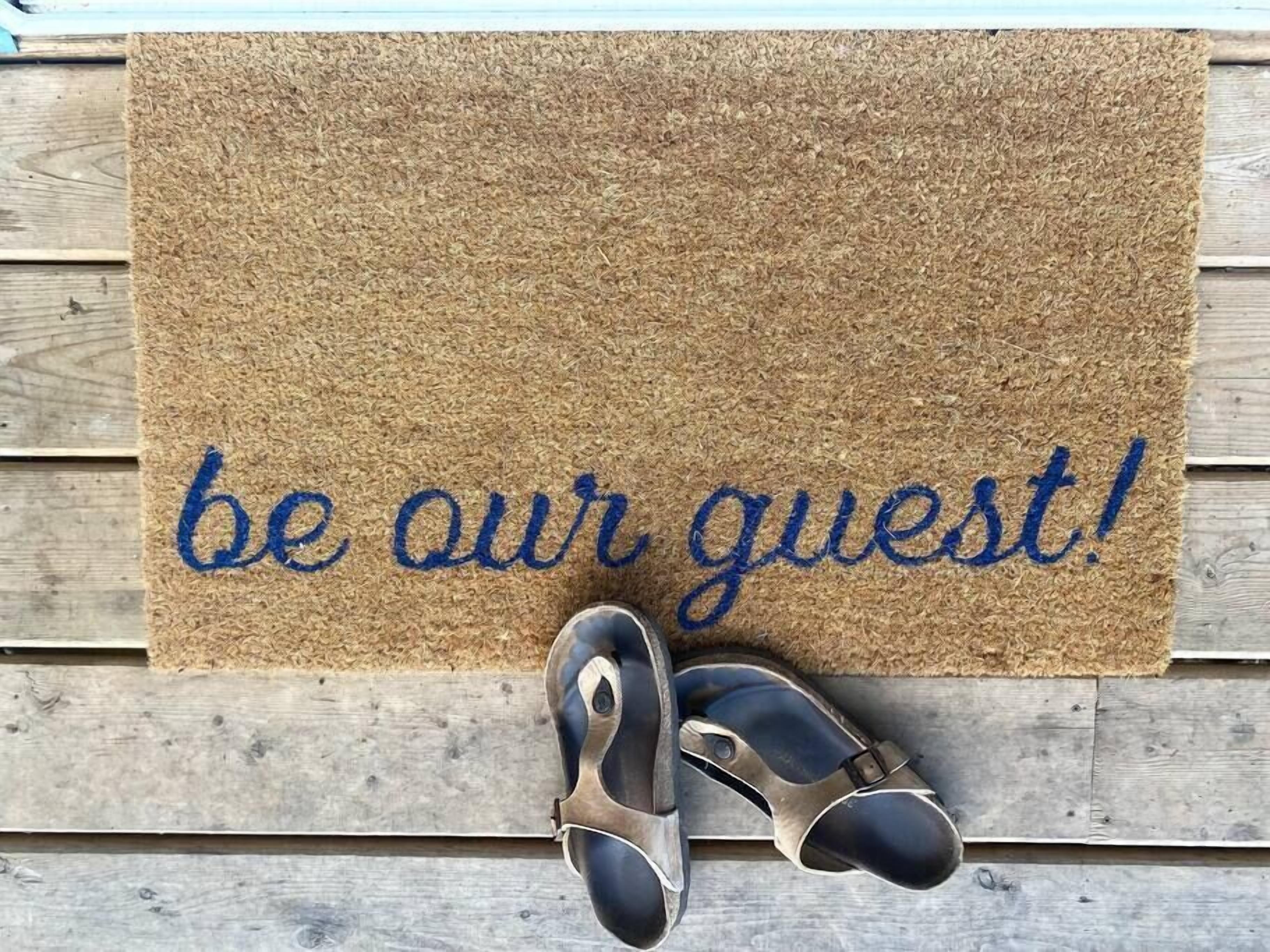 A doormat with the text "be our guest!" and a pair of sandals on a wooden floor.