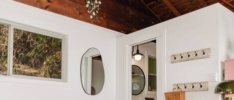A bedroom with a bed, a window, a mirror, a wall-mounted light fixture, and a small bathroom visible through an open door.