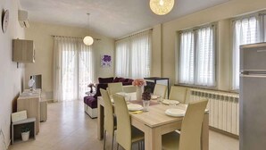 A modern dining area with a wooden table set for four, surrounded by chairs. There is a sofa in the background, a television on a stand, and a radiator under the windows. - Holiday apartment with air conditioning (Vodnjan)