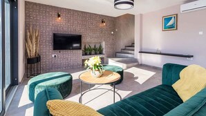 A modern living room with a flat-screen TV, green sofas, a round wooden coffee table, and a staircase.