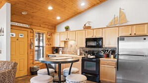 Una cocina con armarios de madera, un refrigerador de acero inoxidable, un microondas y una estufa. Hay una mesa con taburetes y una ventana con persianas.