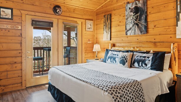 Dormitorio de cabaña de madera con cama, mesita de noche, lámpara, ventana y puerta que da a un balcón.
