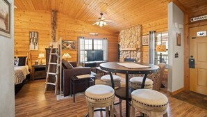 Interior acogedor de una cabaña con paredes de madera, una mesa redonda con taburetes, un televisor y una chimenea.