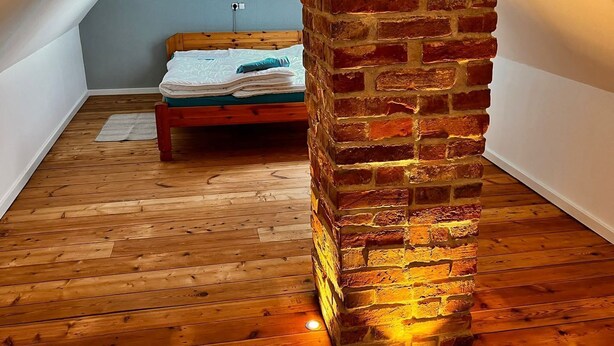 A room with a wooden floor, a bed with a green headboard, and a brick pillar.
