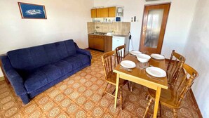 Een knus woongedeelte met een bank, eettafel, stoelen, een kitchenette en vloerbedekking met een patroon.