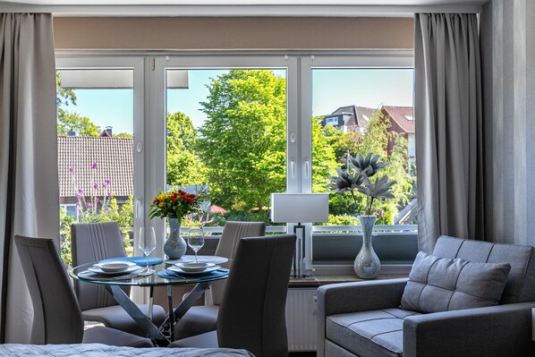 A modern room with a dining table, chairs, a sofa, and large windows with curtains.