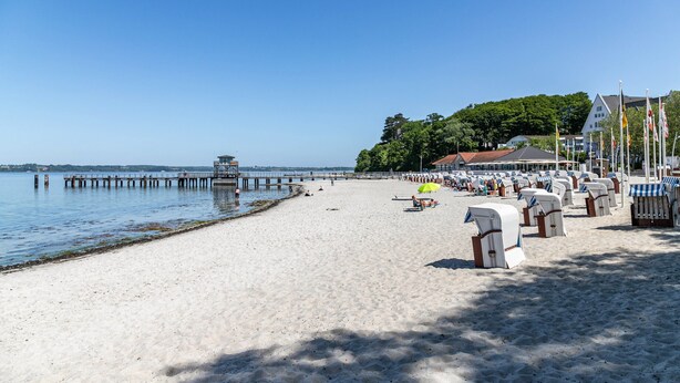 Una playa con un muelle que se adentra en aguas tranquilas, una fila de sillas de playa y un edificio con techo rojo al fondo.