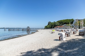 A beach with a pier extending into calm water, a row of beach chairs, and a building with a red roof in the background.