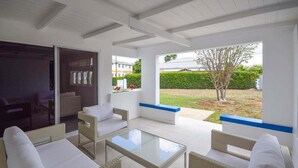 Une terrasse ensoleillée, meublée de meubles en osier, avec une table en verre, et offrant une vue sur un jardin bien entretenu et des bâtiments en arrière-plan.