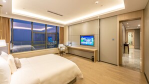 A modern hotel room with a large bed, a desk, a TV, and a view of the city at night.