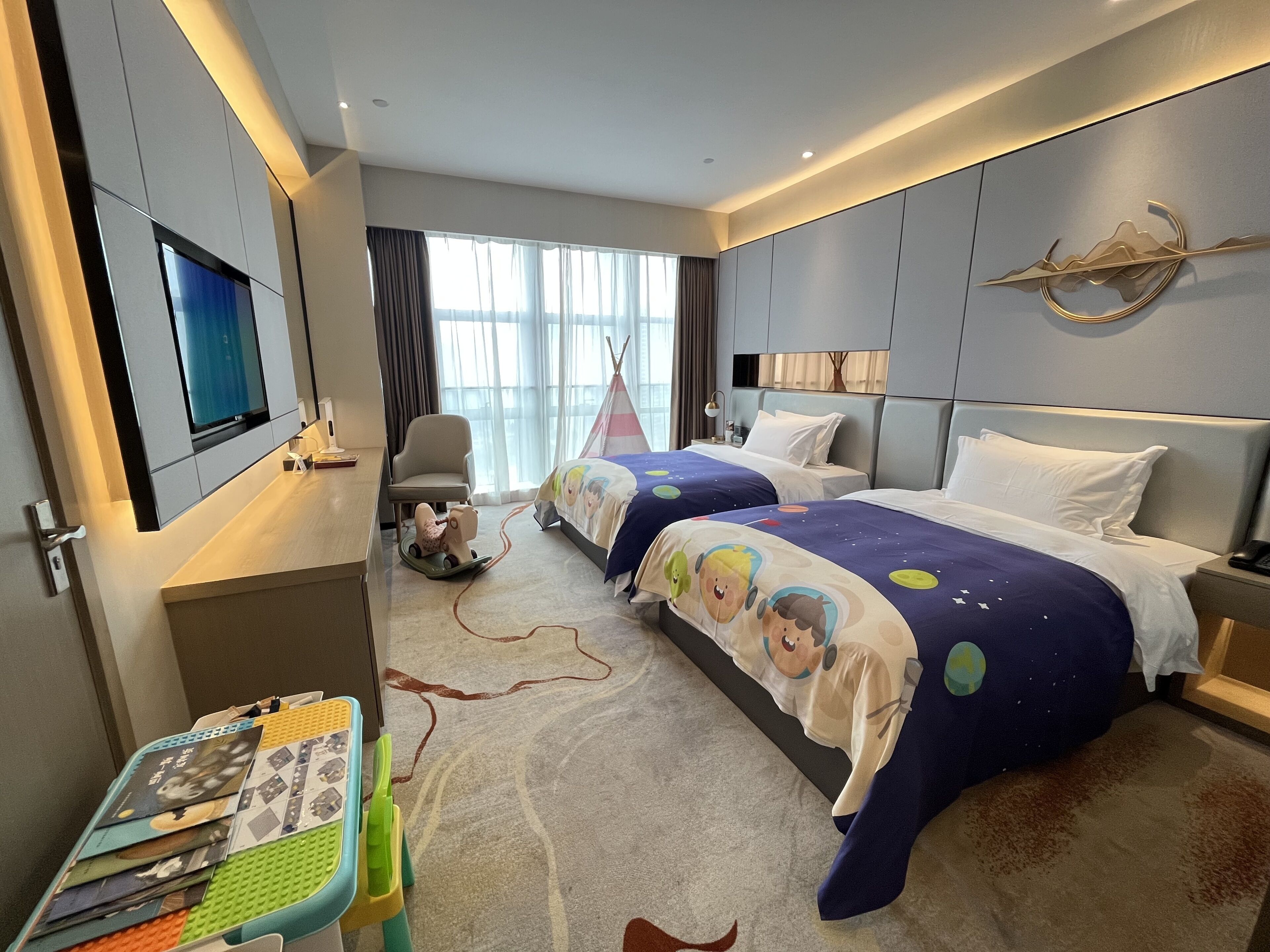 Habitación de hotel con dos camas, un escritorio con una computadora portátil, un televisor y un espacio de juegos para niños.