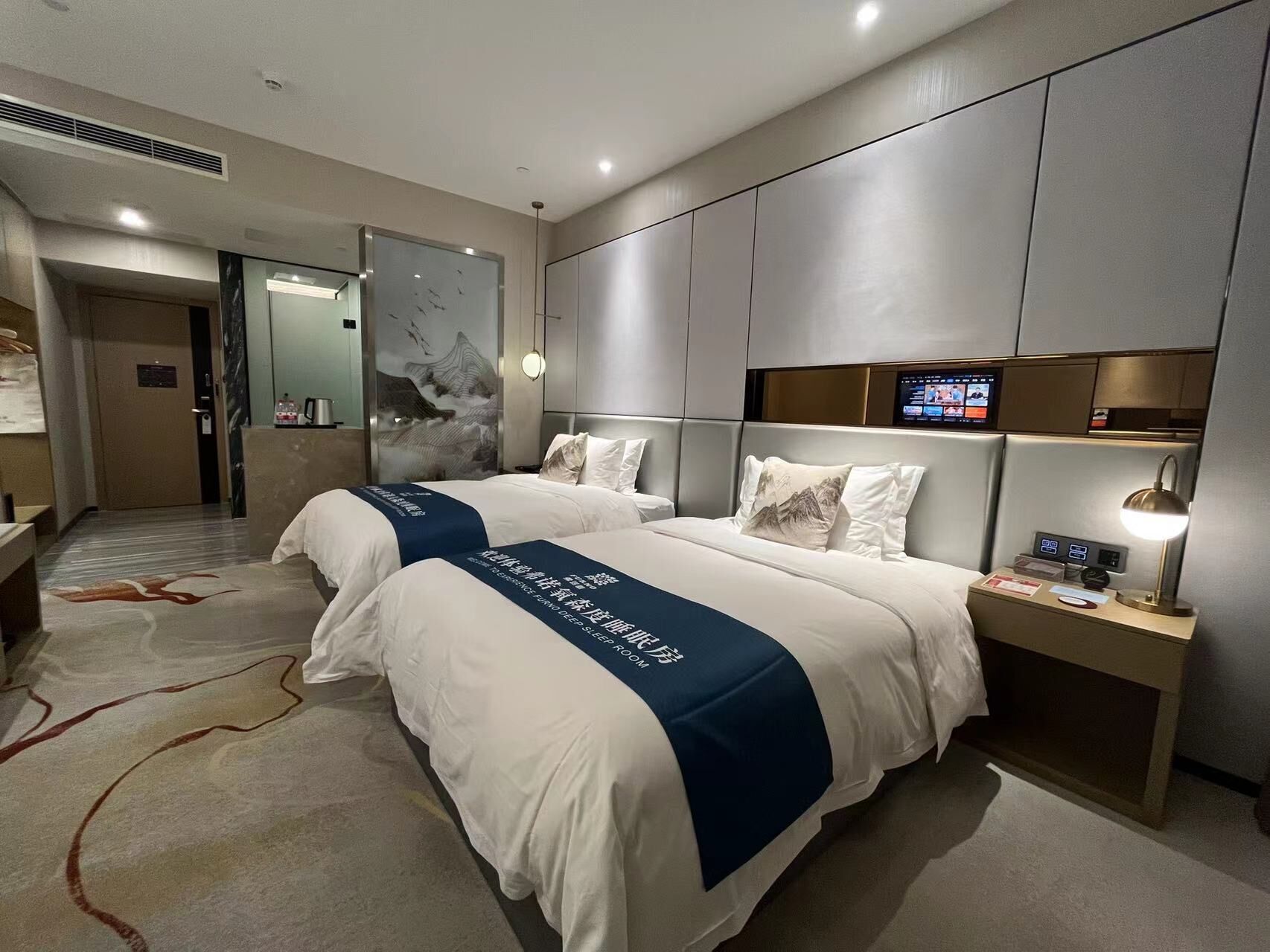 A modern hotel room with two beds, a digital screen, and a bedside table with a lamp.