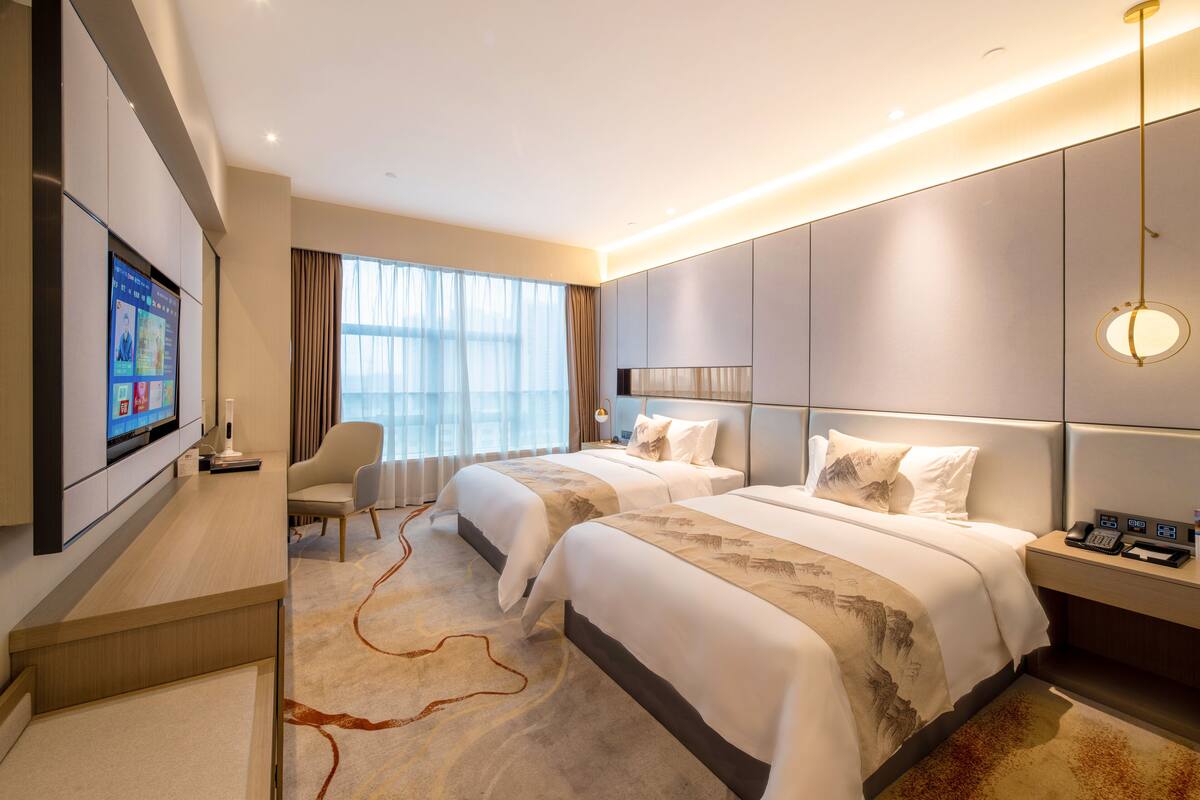 Ein Hotelzimmer mit zwei Betten, einem Fernseher, einem Schreibtisch und einem großen Fenster mit Vorhängen.