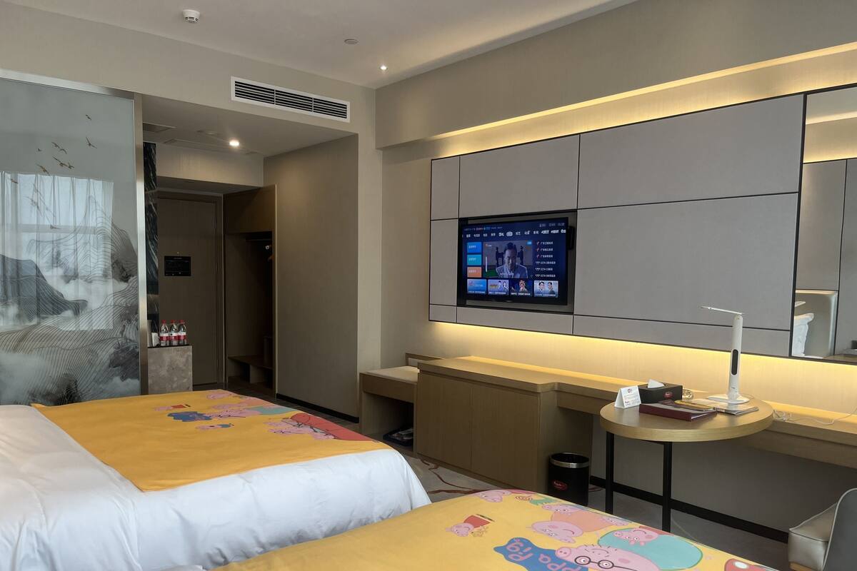 Ein Hotelzimmer mit zwei Betten, einem an der Wand befestigten Fernseher, einem kleinen Tisch mit Lampe und einem Mülleimer.