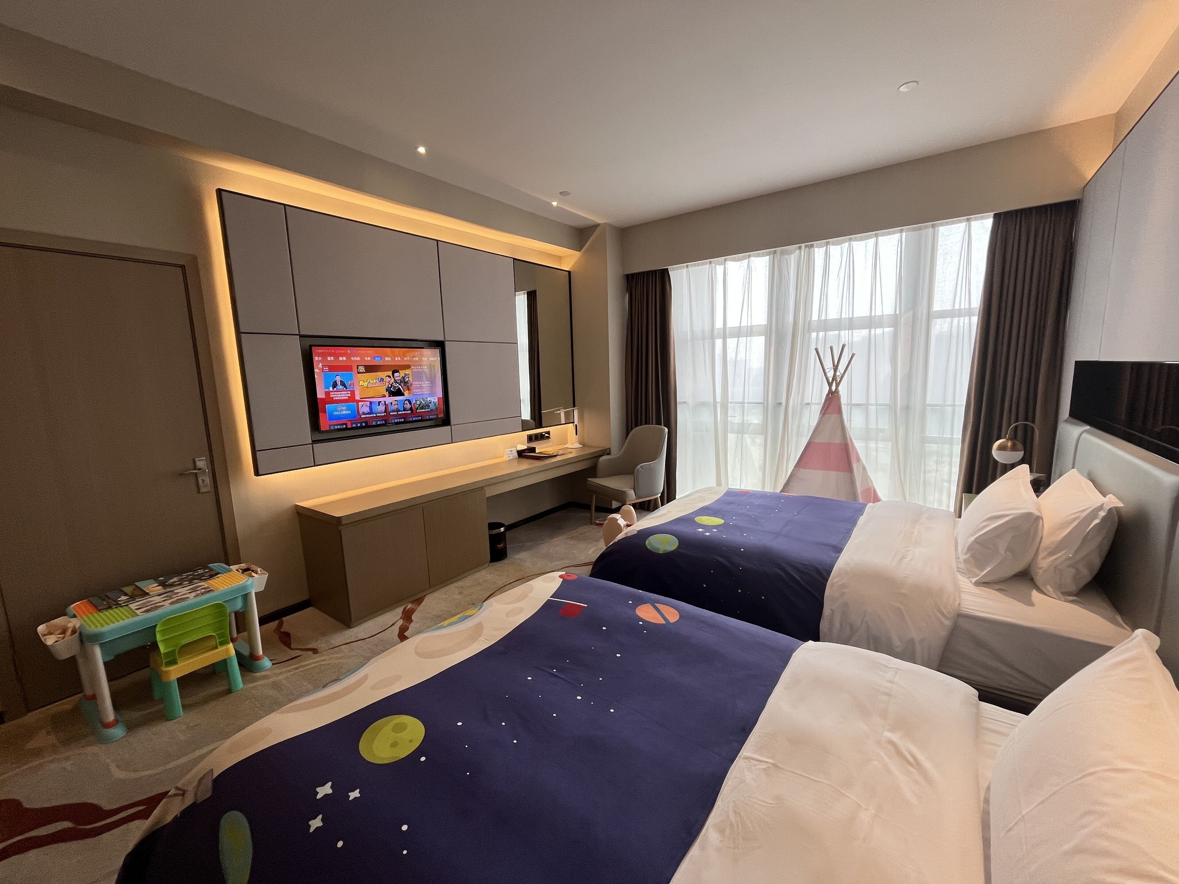 A hotel room with two beds, a desk, a TV, a small table, and a teepee.
