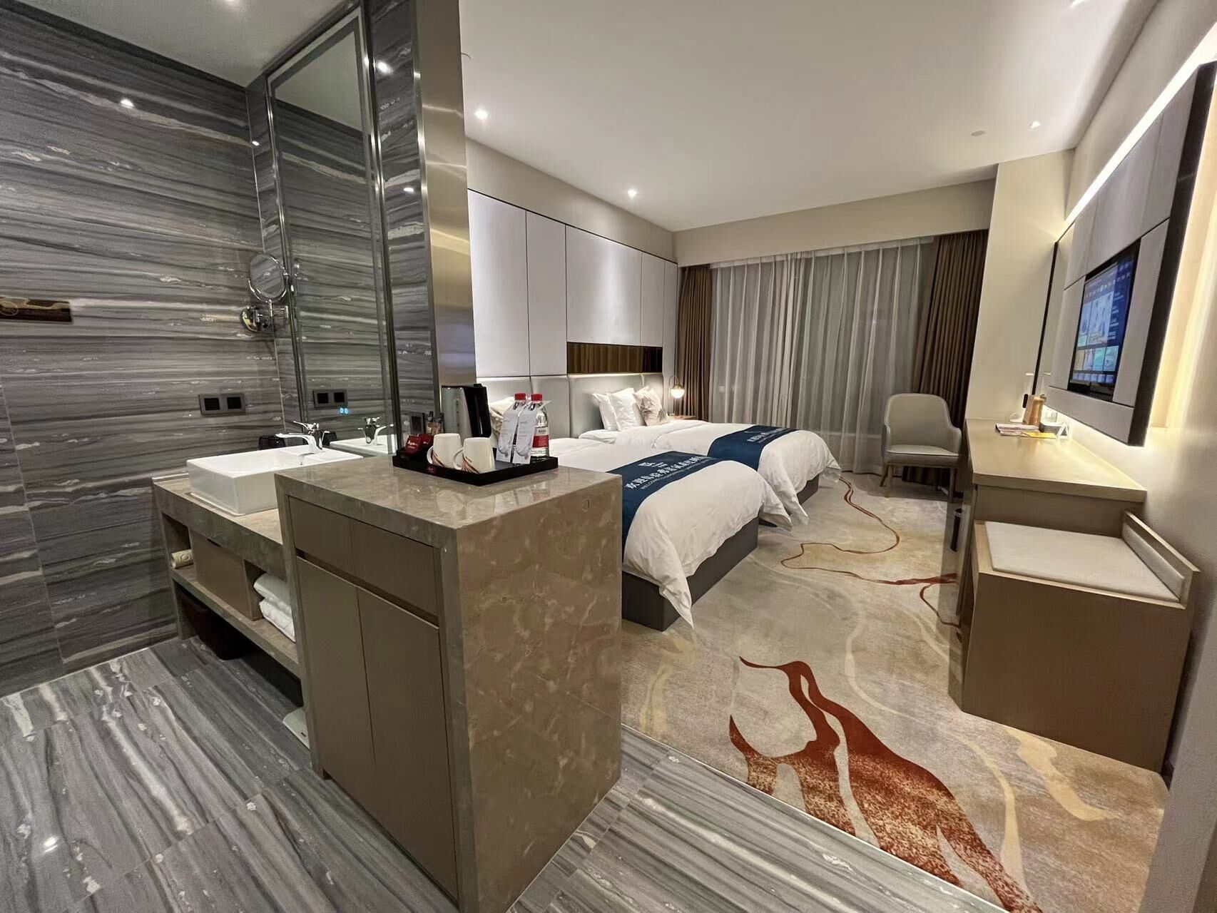 Et moderne hotelværelse med en stor seng, to sengeborde, et skrivebord, en stol og et badeværelse med et stort spejl og vask.