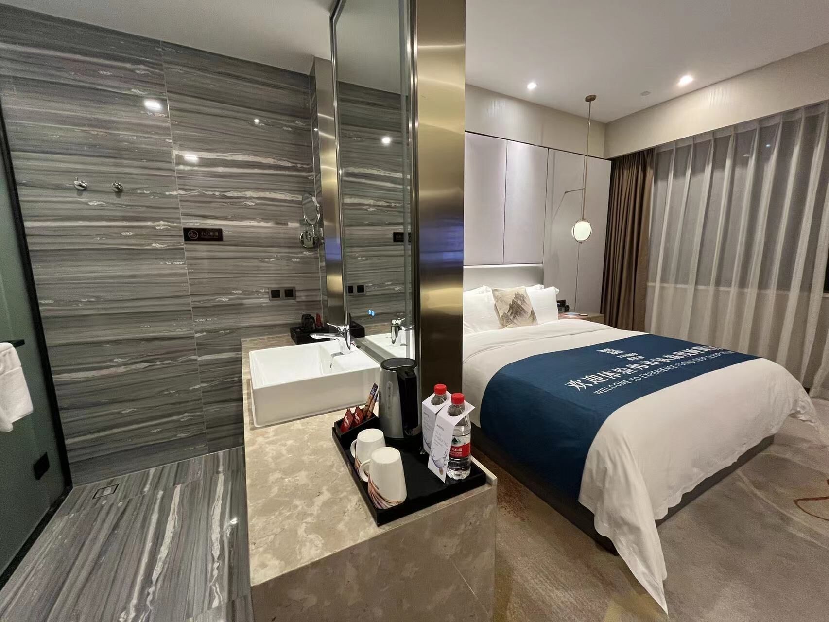 Una habitación de hotel moderna con una pared de mármol, una cama grande con cubrecama azul, un lavamanos blanco y un espejo.