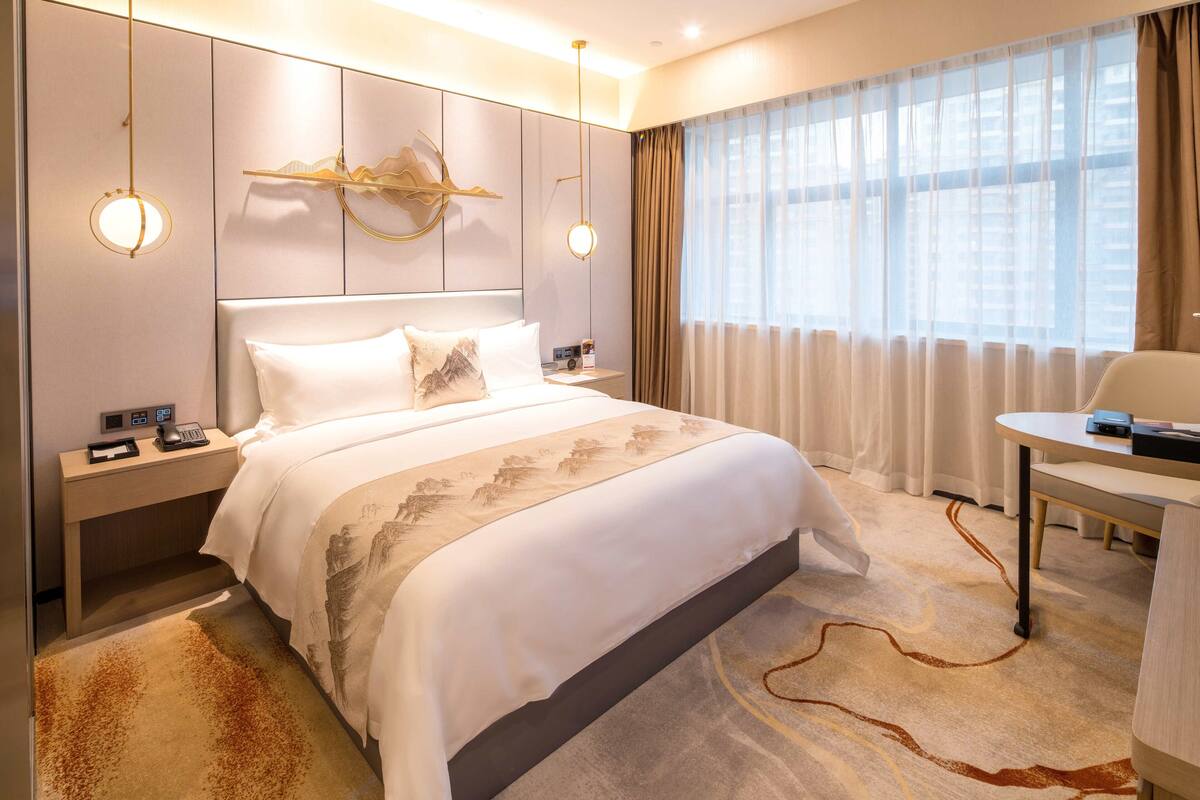 Ein Hotelzimmer mit einem großen Bett, Nachttischen, einem Schreibtisch und Blick auf die Stadt durch transparente Vorhänge.