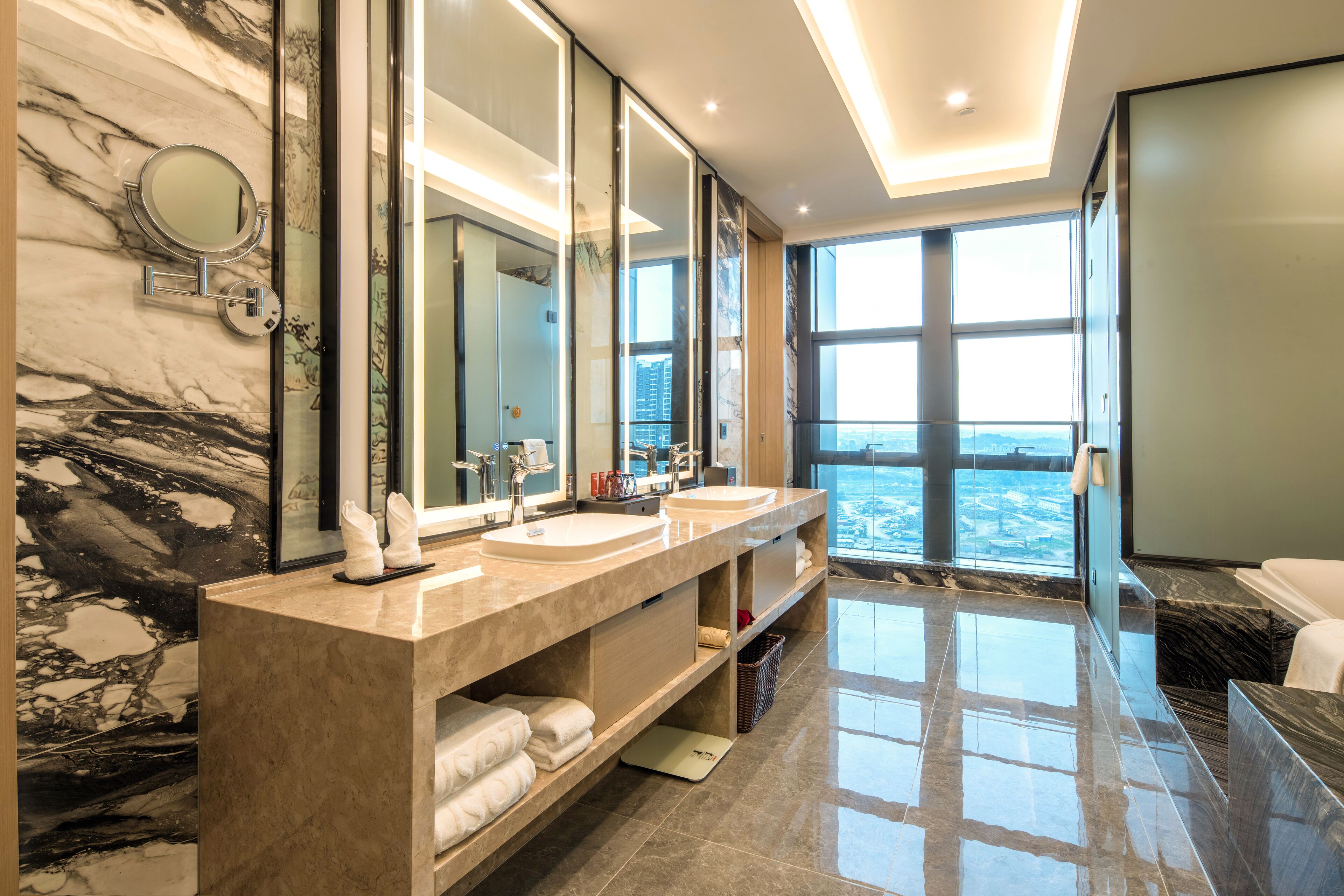 A modern bathroom with a marble backsplash, dual sinks, a large mirror, and a view of the city through large windows.