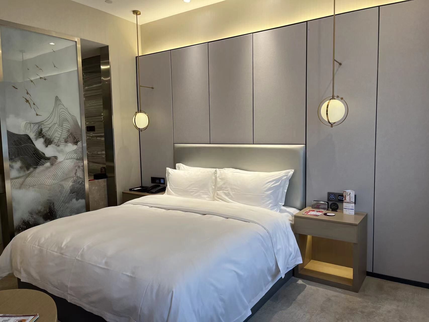 A modern hotel room with a large bed, a bedside table with a lamp, and a wall-mounted mirror.