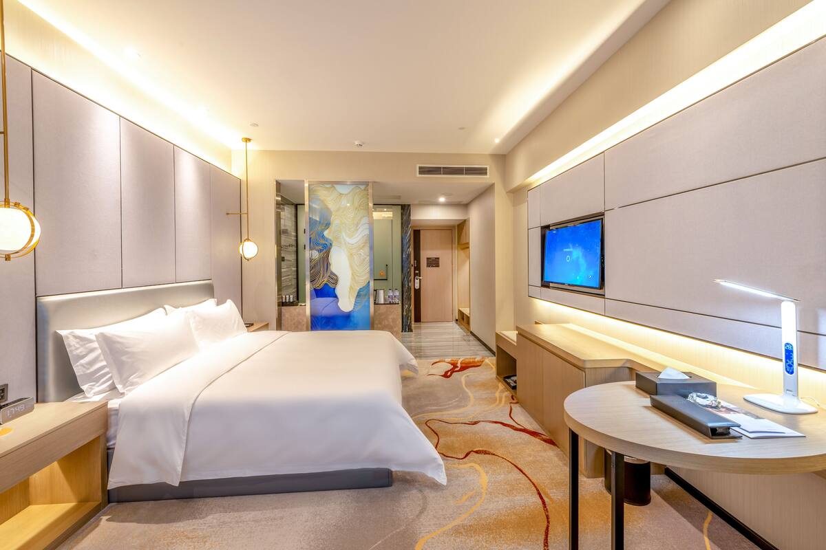 Ein Hotelzimmer mit einem großen Bett, Nachttischen, einem Schreibtisch mit Computer, einem Fernseher und einem Wandbild.