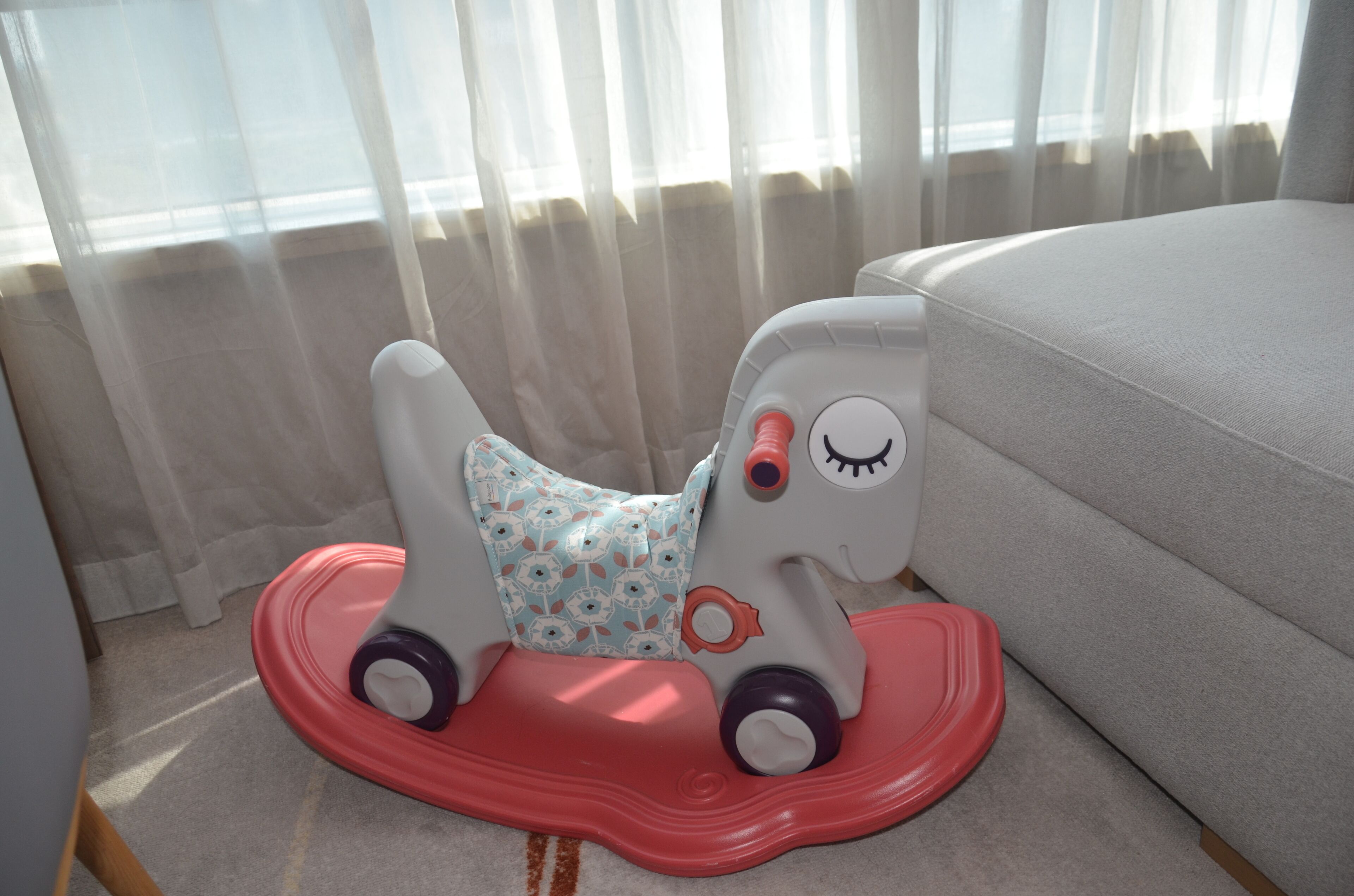A toy horse on a red platform near a window with sheer curtains.