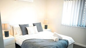 En snyggt bäddad säng med ett rutigt överkast, två sänglampor och ett fönster med persienner.