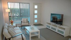 Ett modernt vardagsrum med en vit soffa, ett soffbord, en tv på ett vitt tv-bord och ett fönster med persienner.