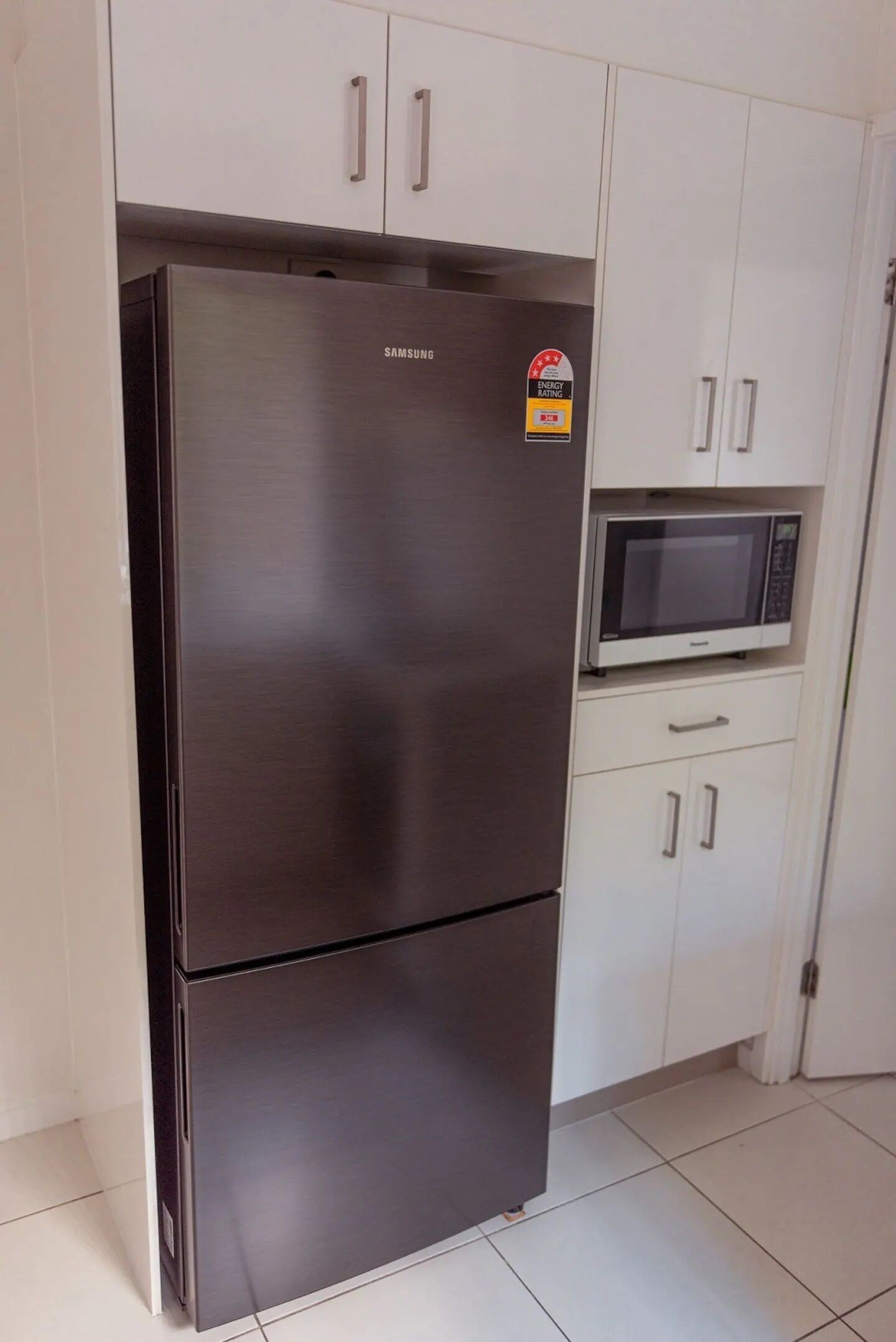 Ein Edelstahl-Kühlschrank von Samsung mit einer Mikrowelle darüber, weißen Schränken und einem gefliesten Boden.