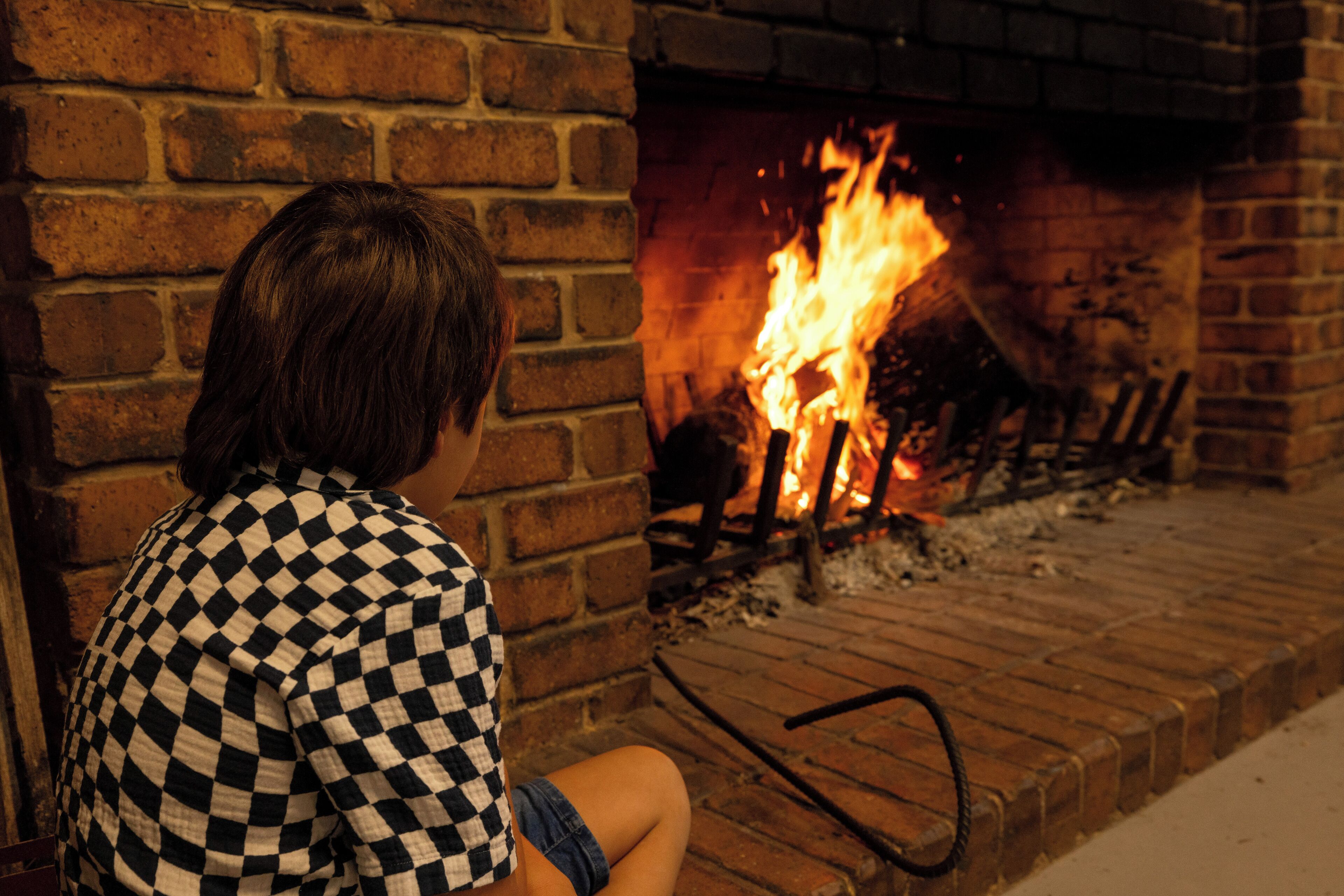 A person sitting by a brick fireplace with a visible fire.
