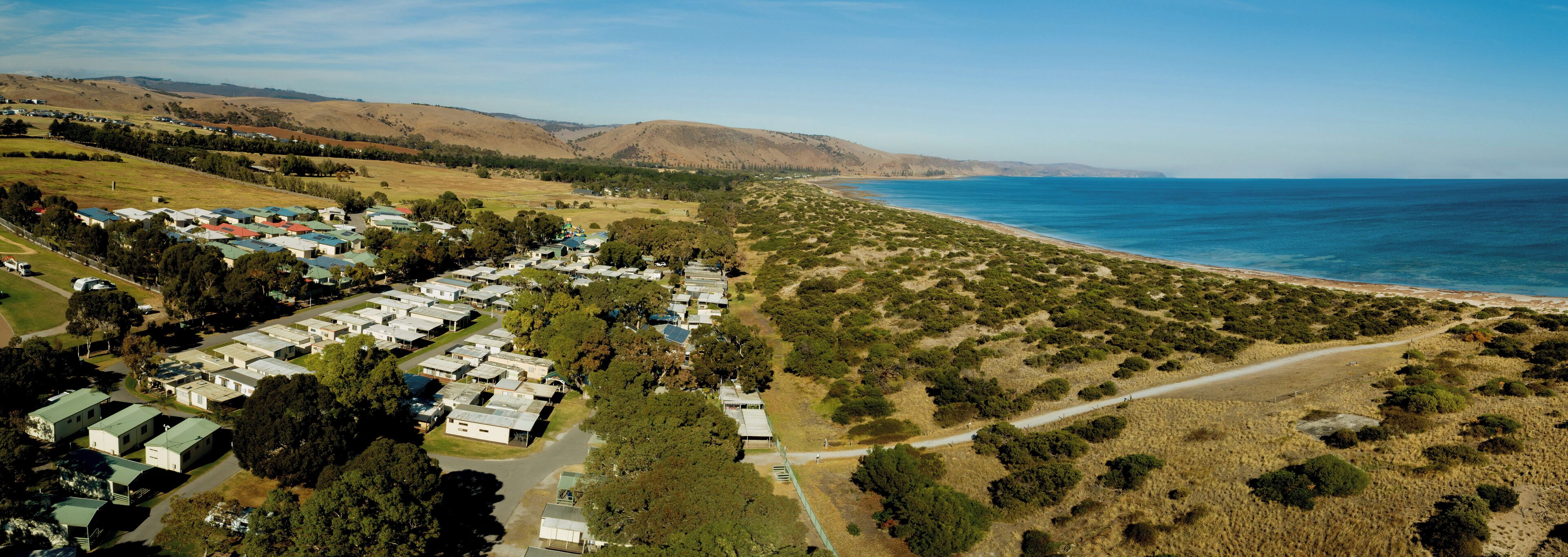 An aerial view of a coastal area with a row of mobile homes along the beach, surrounded by hills and sparse vegetation.