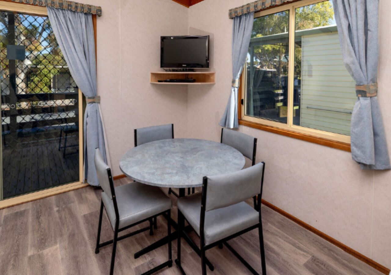 Una mesa redonda con cuatro sillas, un televisor sobre una estantería y una ventana con cortinas.