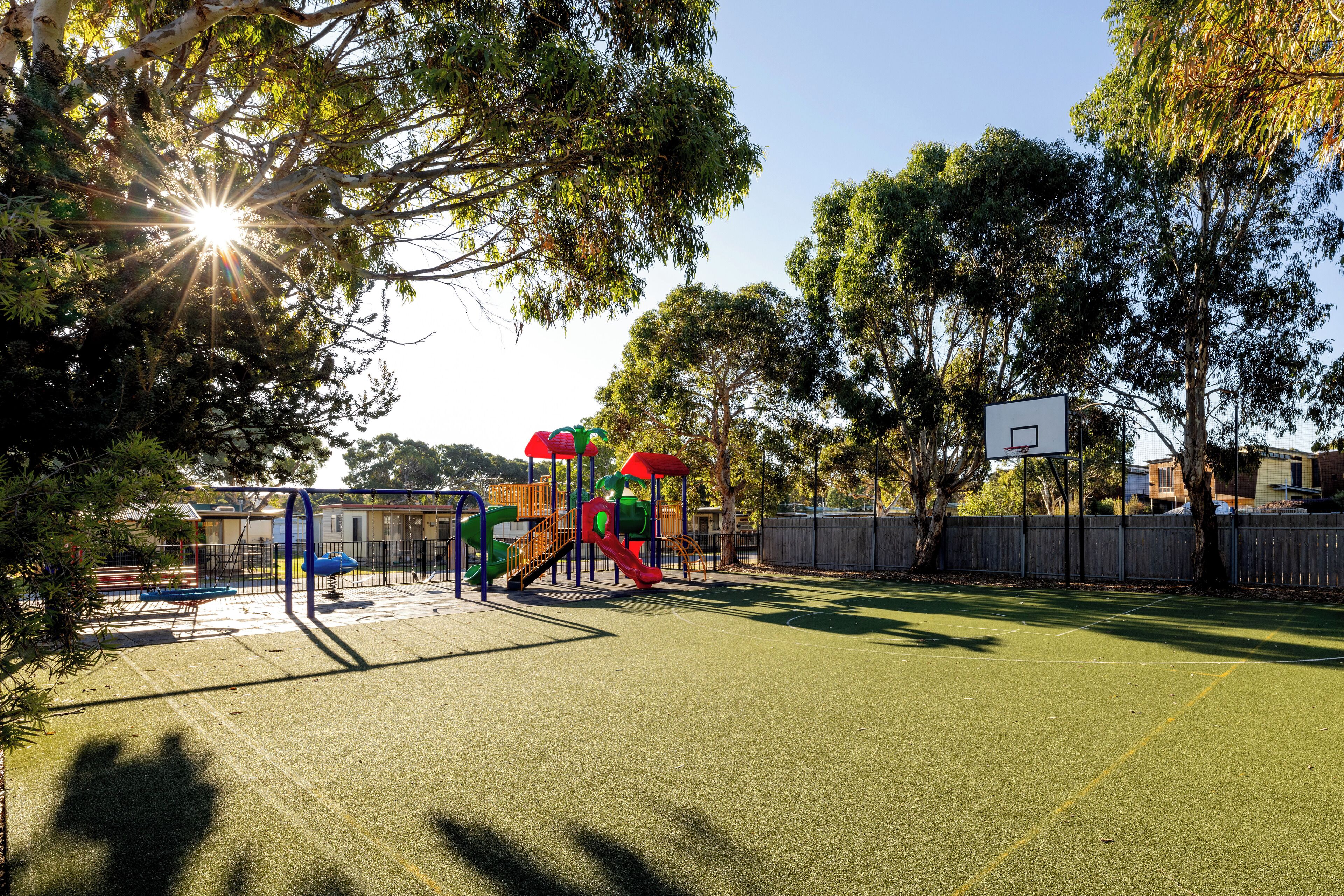 Um parquinho com quadra de basquete, cercado por árvores e uma grade.