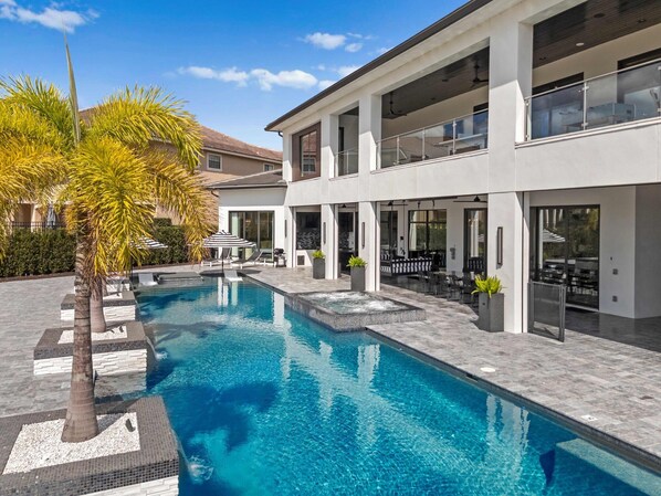 A modern pool area with a clear blue pool, lounge chairs, and a two-story building with large windows and outdoor seating.