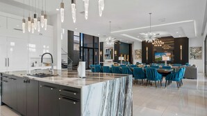 A modern kitchen with a marble island, pendant lights, and a dining area with blue chairs and a chandelier. - Legends Palazzo: Theater, Arcade, Private Pool, Free Water Park, Golf View (Kissimmee)