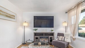 A living room with a fireplace, a flat-screen TV, two floor lamps, a grey sofa, and a glass coffee table. - Cozy Boise Gem: Private Yard, Game Room & Workspace
 (BOISE)