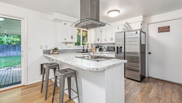 A modern kitchen with white cabinets, a stainless steel refrigerator, and a marble island with bar stools. - Cozy Boise Gem: Private Yard, Game Room & Workspace
 (BOISE)