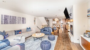 Ein modernes Wohnzimmer mit einer Couch, Hocker und einem Couchtisch. Eine Treppe führt zu einer Küche mit Holzschränken und einem Essbereich.