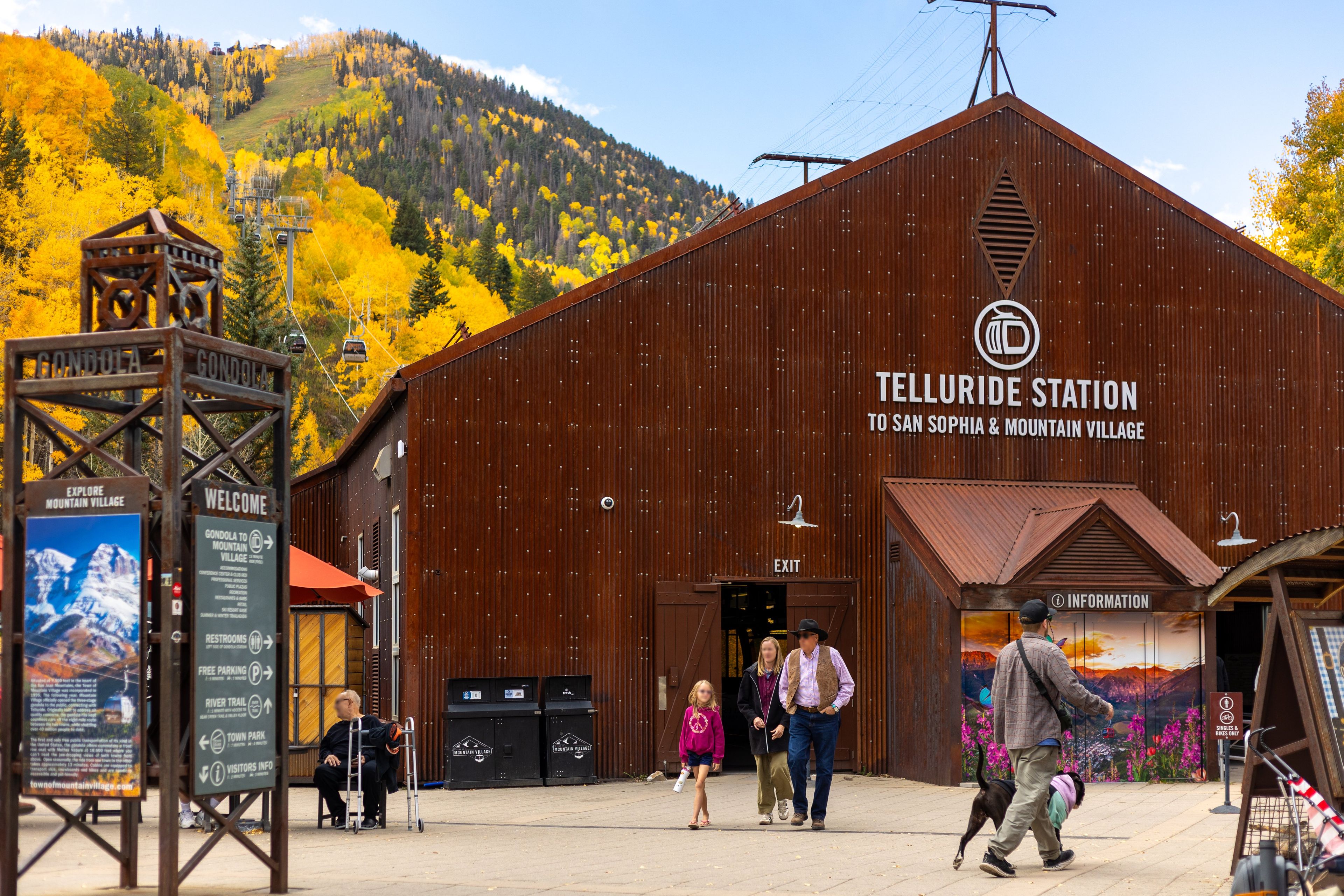 A rustic wooden building labeled 'Telluride Station' with a sign for 'San Sophia & Mountain Village' and a 'Welcome' sign.