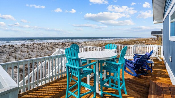 Eine Terrasse mit weißer Brüstung, einem runden Tisch und blauen Stühlen mit Blick auf einen Strand.